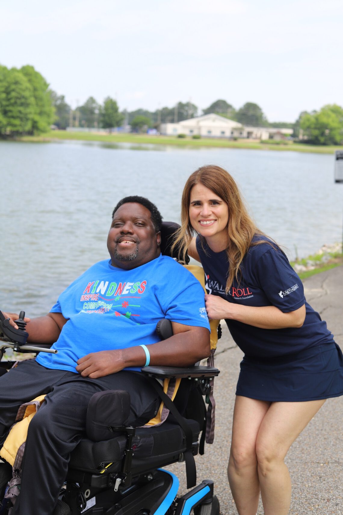 Patient in a wheelchair poses with a therapist while outside on a walk path in front of a pond